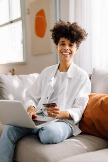 Fröhliche junge Frau, die einen Online-Verkauf zu Hause genießt. Fröhliche junge Frau, die in die Kamera lächelt, während sie an ihrem Laptop einkauft. Junge Frau, die allein auf einer Couch in ihrem Wohnzimmer sitzt. - JLPSF04220
