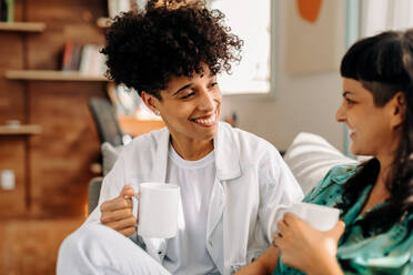 Weibliches Paar beim Kaffee trinken zu Hause. Glückliches junges lesbisches Paar, das sich gegenseitig anlächelt, während es sich in seinem Wohnzimmer entspannt. Junges LGBTQ+ Paar, das Zeit miteinander zu Hause verbringt. - JLPSF04222