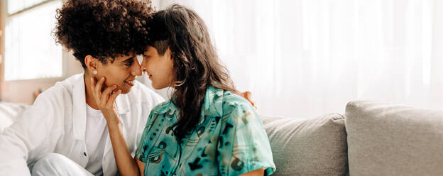 Young female couple sharing an intimate moment at home. Two affectionate female lovers touching their noses together. Romantic young lesbian couple sitting together in their living room. - JLPSF04228