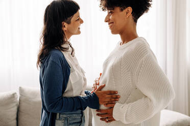 Feeling a surrogate mother's belly bump. Happy young woman smiling while feeling the movement of a pregnant woman's baby. Young woman spending time with her surrogate at home. - JLPSF04235