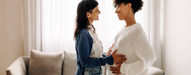 Smiling woman feeling a pregnant woman's belly. Happy young woman feeling the movement of a pregnant woman's baby. Cheerful young woman spending time with her surrogate at home. - JLPSF04236