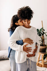 Romantic young woman embracing her pregnant wife at home. Affectionate young woman kissing her wife's neck while embracing her belly bump. Pregnant lesbian couple bonding fondly at home. - JLPSF04244