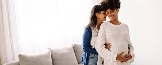 Expectant lesbian couple bonding at home. Happy pregnant woman smiling cheerfully while standing with her wife indoors. Pregnant lesbian couple spending quality time together. - JLPSF04248