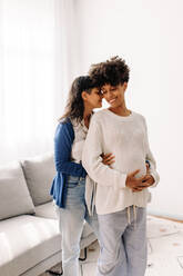 Queer couple expecting a baby. Happy young pregnant woman smiling cheerfully while standing with her wife indoors. Pregnant lesbian couple spending quality time together at home. - JLPSF04249