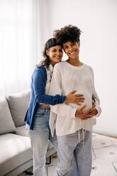 Ein schwangeres lesbisches Paar lächelt in die Kamera, während es in einem Haus steht. Eine glückliche junge Frau umarmt ihre schwangere Frau von hinten. Ein schwangeres lesbisches Paar freut sich auf ihr ungeborenes Baby. - JLPSF04250