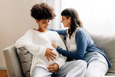 Female parents-to-be sitting together at home. Happy young woman smiling cheerfully while embracing her pregnant wife. Pregnant lesbian couple spending quality time together. - JLPSF04252