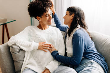 Queer parents-to-be sitting together at home. Happy young pregnant couple smiling at each other in their living room. Pregnant lesbian couple spending quality time together. - JLPSF04253