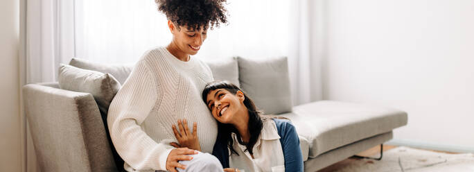 Lesbian mother-to-be listening to her unborn baby kicking. Happy young woman smiling cheerfully while listening to her wife's pregnant belly at home. Young lesbian couple expecting a baby. - JLPSF04256