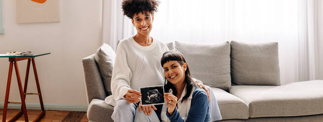 Lesbische werdende Mütter halten ihr Ultraschallbild hoch. Ein schwangeres lesbisches Paar lächelt in die Kamera, während es ein Ultraschallbild seines ungeborenen Babys hält. Ein junges weibliches Paar erwartet ein Baby. - JLPSF04259