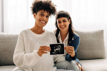 Ein verheiratetes lesbisches Paar hält ein Ultraschallbild ihres ungeborenen Babys hoch. Ein schwangeres lesbisches Paar lächelt in die Kamera, während es sein Ultraschallbild hält. Ein junges queeres Paar erwartet ein Baby. - JLPSF04260