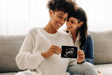 Gleichgeschlechtliches schwangeres Paar, das sein Ultraschallbild hochhält. Ein schwangeres lesbisches Paar, das fröhlich lächelt, während es ein Ultraschallbild seines ungeborenen Babys in der Hand hält. Junges queeres Paar, das ein Baby erwartet. - JLPSF04261