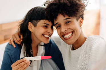 Weibliches Paar macht ein Selfie mit ihrem positiven Schwangerschaftstest. Glückliches junges weibliches Paar lächelt fröhlich, nachdem es einen Schwangerschaftstest zu Hause gemacht hat. Junges lesbisches Paar, das ein Baby erwartet. - JLPSF04263