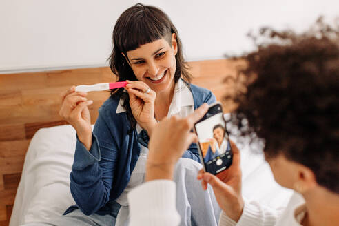 Cheerful young woman posing for a picture holding her home pregnancy test. Happy young female couple recording their pregnancy test results for their blog. Young lesbian couple expecting a baby. - JLPSF04265