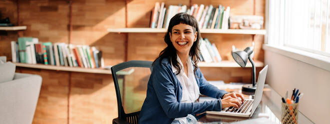 Businesswoman smiling while working from home. Successful young businesswoman looking away while typing on a laptop at her home office desk. Happy young woman working from home during quarantine. - JLPSF04275