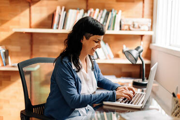 Lächelnde Frau, die in ihrem Büro zu Hause arbeitet. Erfolgreiche junge Geschäftsfrau, die auf einem Laptop tippt, während sie in ihrem Büro zu Hause sitzt. Glückliche junge Frau, die während der Quarantäne von zu Hause aus arbeitet. - JLPSF04276
