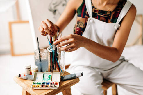 Unrecognizable female painter picking a brush from a jar. Anonymous young female painter preparing to start drawing a new painting in her studio. Young woman sitting alone in her art studio. - JLPSF04295