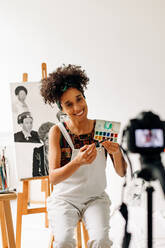 Bloggerin, die aus ihrem Atelier live streamt. Glückliche junge Malerin, die lächelt, während sie eine Malanleitung mit einer Kamera aufnimmt. Junge Freiberuflerin, die in ihrem Kunstatelier einen Videoblog erstellt. - JLPSF04298