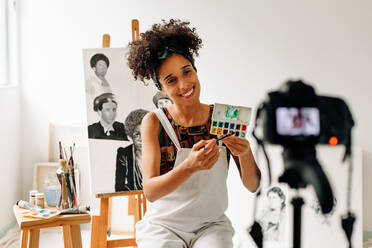 Malerin, die in ihrem Atelier einen Kunstlehrgang aufnimmt. Glückliche junge Malerin, die lächelt, während sie mit einer Kamera live streamt. Junge Freiberuflerin, die in ihrem Atelier einen Videoblog erstellt. - JLPSF04299