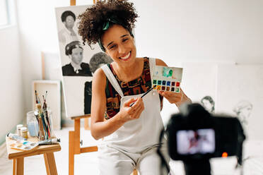 Freiberuflerin, die in ihrem Atelier einen Kunstlehrgang aufnimmt. Glückliche junge Malerin, die lächelt, während sie mit einer Kamera live streamt. Junge Malerin, die in ihrem Atelier einen Videoblog erstellt. - JLPSF04300