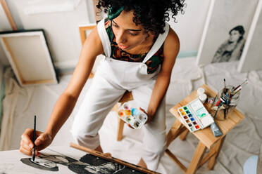 Top view of a young female painter working on a new project. Focused young female painter drawing on a canvas in her art studio. Creative young woman working alone in a workshop. - JLPSF04301