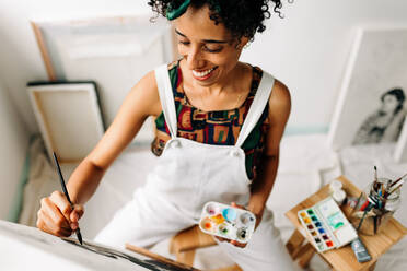 Glückliche junge Malerin bei der Arbeit in einem Atelier. Junge Malerin lächelt fröhlich, während sie in ihrem Atelier auf eine Leinwand zeichnet. Kreative junge Frau bei der Arbeit an einem neuen Projekt. - JLPSF04303