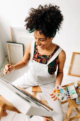 Fröhliche Malerin, die in einem Atelier an einem neuen Projekt arbeitet. Junge Malerin, die fröhlich lächelt, während sie in ihrem Atelier auf eine Leinwand malt. Kreative junge Frau, die allein arbeitet. - JLPSF04304