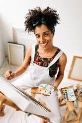 Selbstbewusste junge Malerin, die auf einer Leinwand zeichnet. Junge Malerin, die in die Kamera lächelt, während sie in ihrem Atelier an einem neuen Projekt arbeitet. Kreative junge Frau, die in einer Werkstatt arbeitet. - JLPSF04305