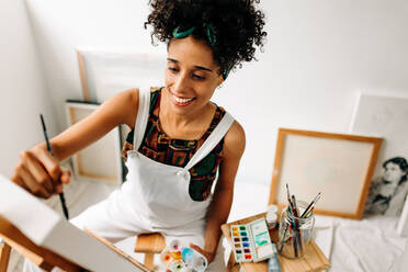 Kreative junge Frau, die fröhlich lächelt, während sie in ihrem Atelier an einem neuen Projekt arbeitet. Glückliche junge Malerin bei der Arbeit in einer Werkstatt. - JLPSF04306