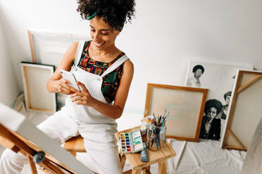 Vloggerin, die ihr Kunstwerk in ihrer Werkstatt fotografiert. Glückliche junge Malerin, die lächelt, während sie ihre bemalte Leinwand mit einem Smartphone festhält. Freiberuflerin, die einen Online-Malblog erstellt. - JLPSF04309