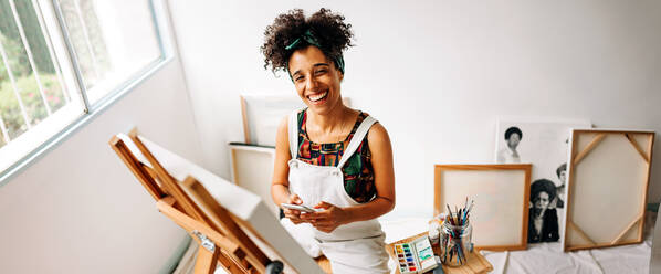 Fröhliche Malerin, die in ihrem Atelier ein Smartphone in der Hand hält. Glückliche junge Malerin, die in die Kamera lächelt, während sie vor ihrer Leinwand sitzt. Junge Frau, die ihren Malerei-Blog online aktualisiert. - JLPSF04310