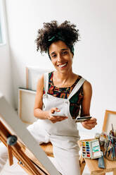 Glückliche junge Malerin, die mit ihrer Kreditkarte und ihrem Smartphone online einkauft. Junge Malerin, die in die Kamera lächelt, während sie vor ihrer Leinwand sitzt. - JLPSF04311