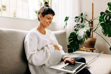 Happy pregnant woman video calling her friends on a laptop. Young pregnant woman smiling cheerfully while speaking with her friends on a video call. Young woman expecting a baby. - JLPSF04312