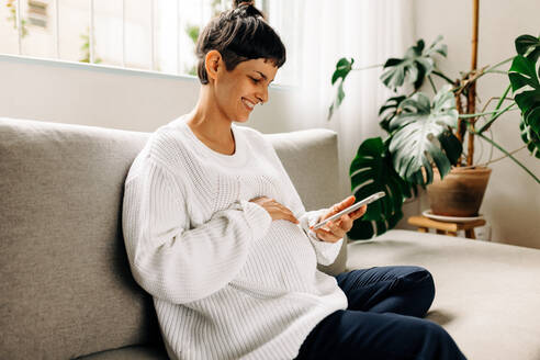 Carefree pregnant woman video calling her friends at home. Happy young pregnant woman smiling cheerfully while speaking with her friends on a video call. Young woman expecting a baby. - JLPSF04313
