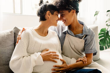 Married lesbian couple expecting a baby. Pregnant lesbian couple smiling and embracing each other while sitting in their living room. Expectant female couple bonding fondly at home. - JLPSF04314