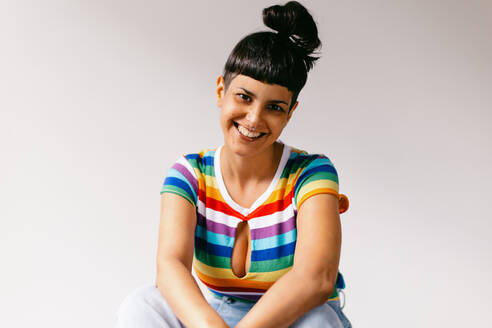 Queer woman celebrating gay pride in a studio. Happy young lesbian woman smiling at the camera against a studio background. Young woman wearing a tank top with LGBTQ+ rainbow colours. - JLPSF04319