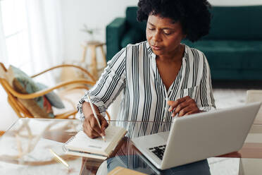 Frau sitzt am Tisch und macht sich Notizen im Heimbüro. Afrikanische Frau arbeitet von zu Hause aus. - JLPSF04321