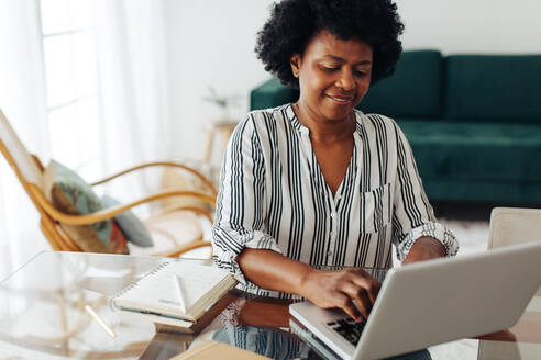 Reife afrikanische Frau, die von zu Hause aus arbeitet. Reife Frau, die an einem Tisch sitzt und einen Laptop im Heimbüro benutzt. - JLPSF04322