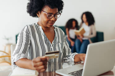 Ältere Frau macht Online-Zahlung auf Laptop mit Familie im Hintergrund. Frau mit Kreditkarte für Online-Zahlung auf Laptop zu Hause. - JLPSF04326