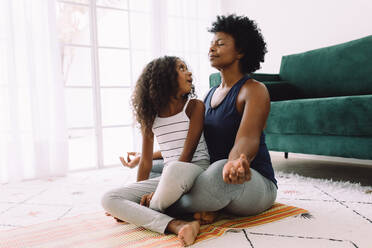 Mädchen sitzt im Schoß der Mutter, während sie zu Hause meditiert. Frau macht Yoga-Meditation mit ihrer Tochter auf dem Schoß. - JLPSF04332