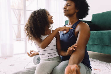 Daughter playing with mother doing yoga meditation in living room. Mature mother doing meditation while her daughter distracting her. - JLPSF04333