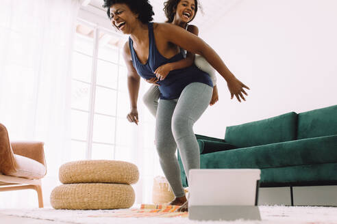 Woman and daughter having fun together while exercising. Mature mother in fitness wear with her daughter on her back while doing online workout at home. - JLPSF04335