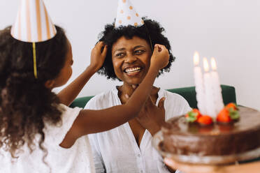 Girl putting party hat mother's head. Woman celebration her birthday with daughter at home. - JLPSF04354