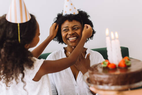 Mädchen setzt Partyhut auf den Kopf der Mutter. Frau feiert ihren Geburtstag mit Tochter zu Hause. - JLPSF04354