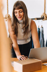 Happy young businesswoman using a laptop while preparing online orders in a clothing store. Young female entrepreneur running an e-commerce small business with her colleague. - JLPSF04360