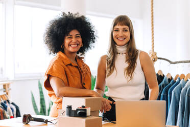 Erfolgreiche Geschäftsfrauen, die in ihrem Secondhand-Laden in die Kamera lächeln. Glückliche Online-Shop-Besitzerinnen, die Pakete für den Versand vorbereiten. Zwei Unternehmerinnen, die ein E-Commerce-Kleinunternehmen betreiben. - JLPSF04361