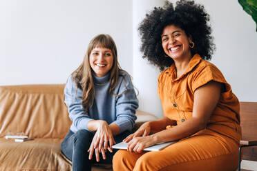 Two successful businesswomen smiling at the camera while sitting together in an office lobby. Two happy female entrepreneurs working as a team in a modern workplace. - JLPSF04368