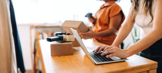 Angestellte eines Online-Shops, die mit ihrem Kollegen an einem Laptop arbeitet. Zwei junge Geschäftsfrauen, die Online-Bestellungen für den Versand vorbereiten. Unternehmerinnen, die ein kleines E-Commerce-Unternehmen betreiben. - JLPSF04376