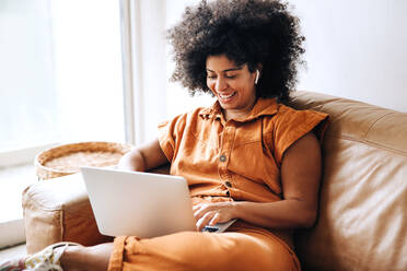 Junge Geschäftsfrau lächelt fröhlich, während sie an einem Laptop in einer Büro-Lobby arbeitet. Glückliche junge Geschäftsfrau sitzt auf einer Couch in einem modernen Arbeitsplatz. - JLPSF04388