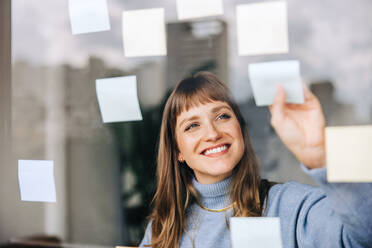 Fröhliche junge Geschäftsfrau, die in einem modernen Büro Haftnotizen an eine Glaswand klebt. Zufriedene Unternehmerin, die ihre Ideen ausarbeitet, während sie an einer neuen kreativen Strategie arbeitet. - JLPSF04402