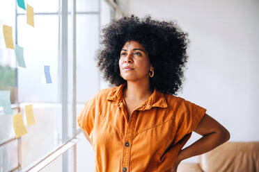 Nachdenkliche junge Geschäftsfrau, die neben einer Glaswand in einem Büro steht und den Blick abwendet. Schwarze Unternehmerin, die ihre kreativen Ideen mit Haftnotizen organisiert. - JLPSF04406